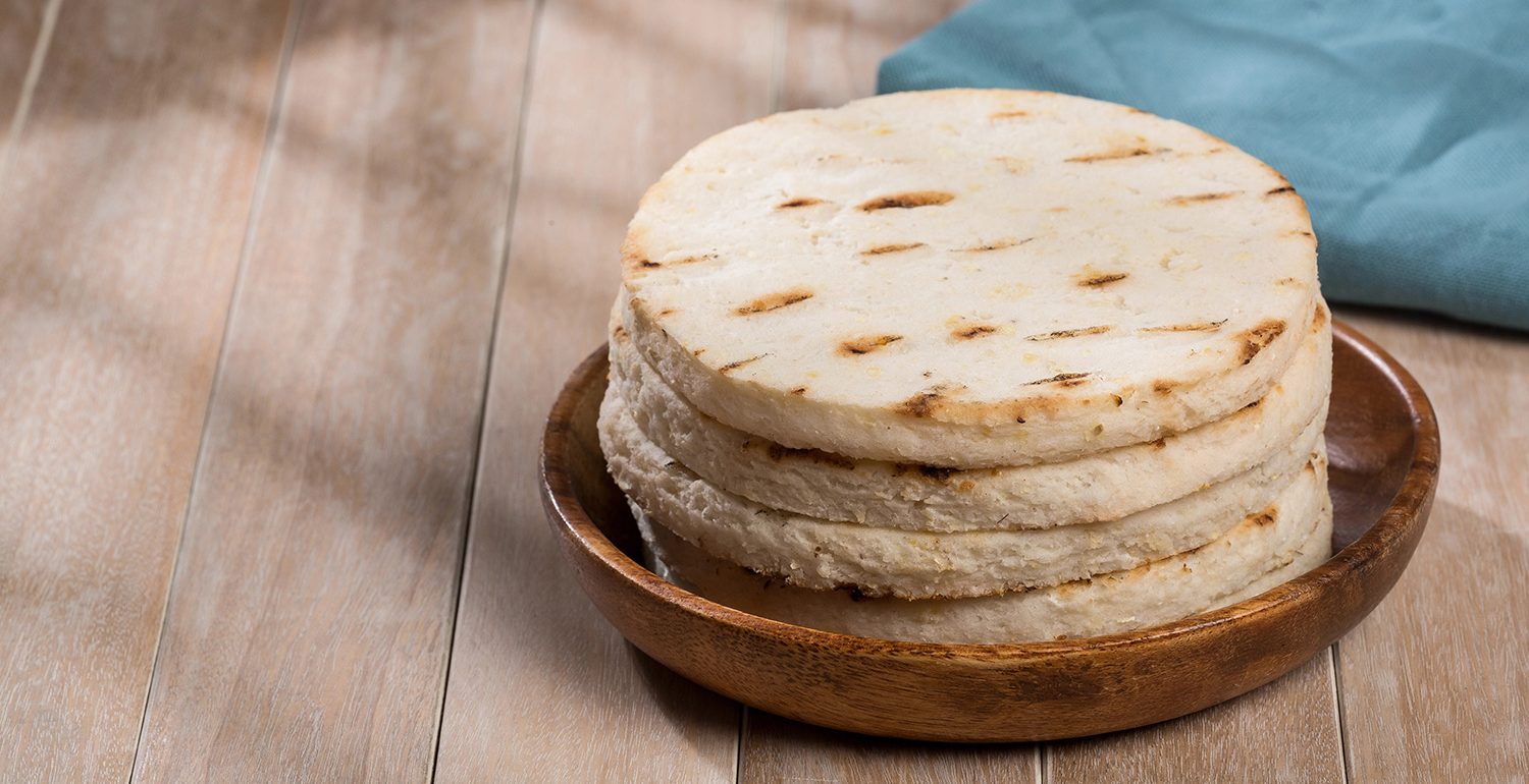 Torre de arepas de maíz asadas, servidas en un plato de madera. Las acompaña una servilleta de tela azul sobre una mesa de madera.